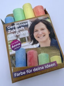 Schachtel mit bunter Straßenmalkreide, Banderole mit Anne Fiedler und den Sätzen „Gemeinsam Zukunft gestalten.“ und „Farbe für deine Ideen.“